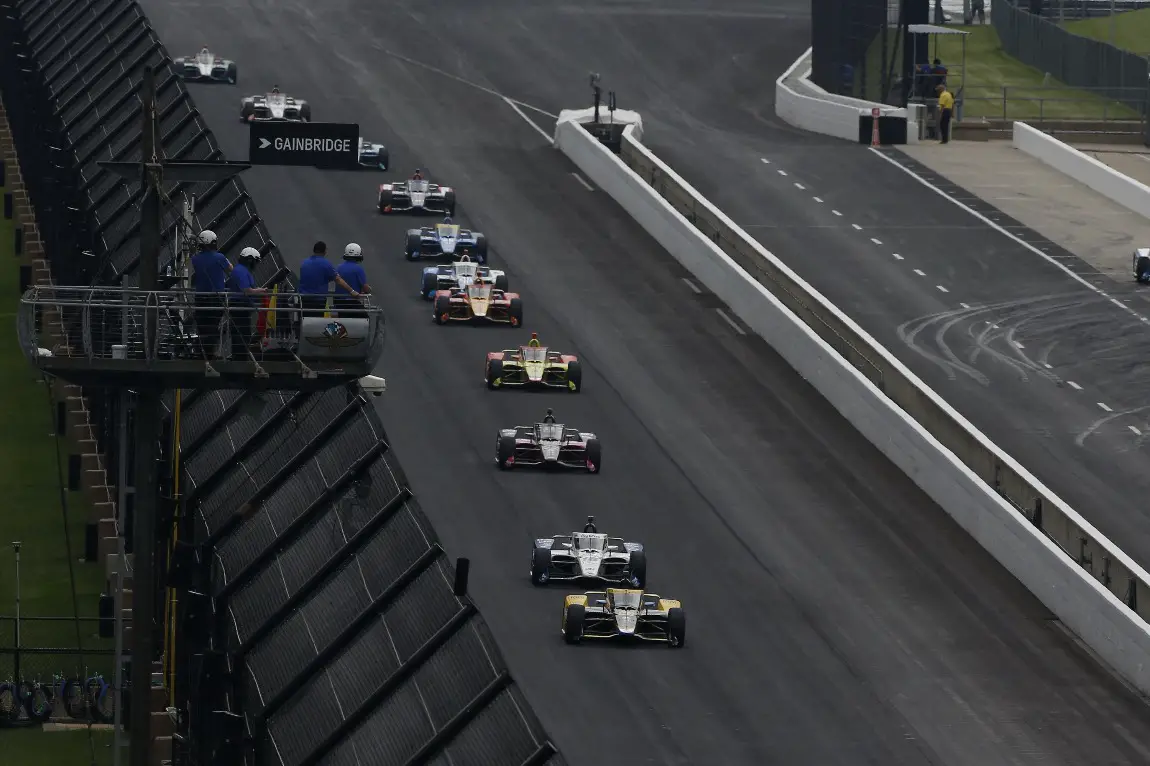 A pack of NTT IndyCar Series drivers driving around the Indianapolis Motor Speedway.