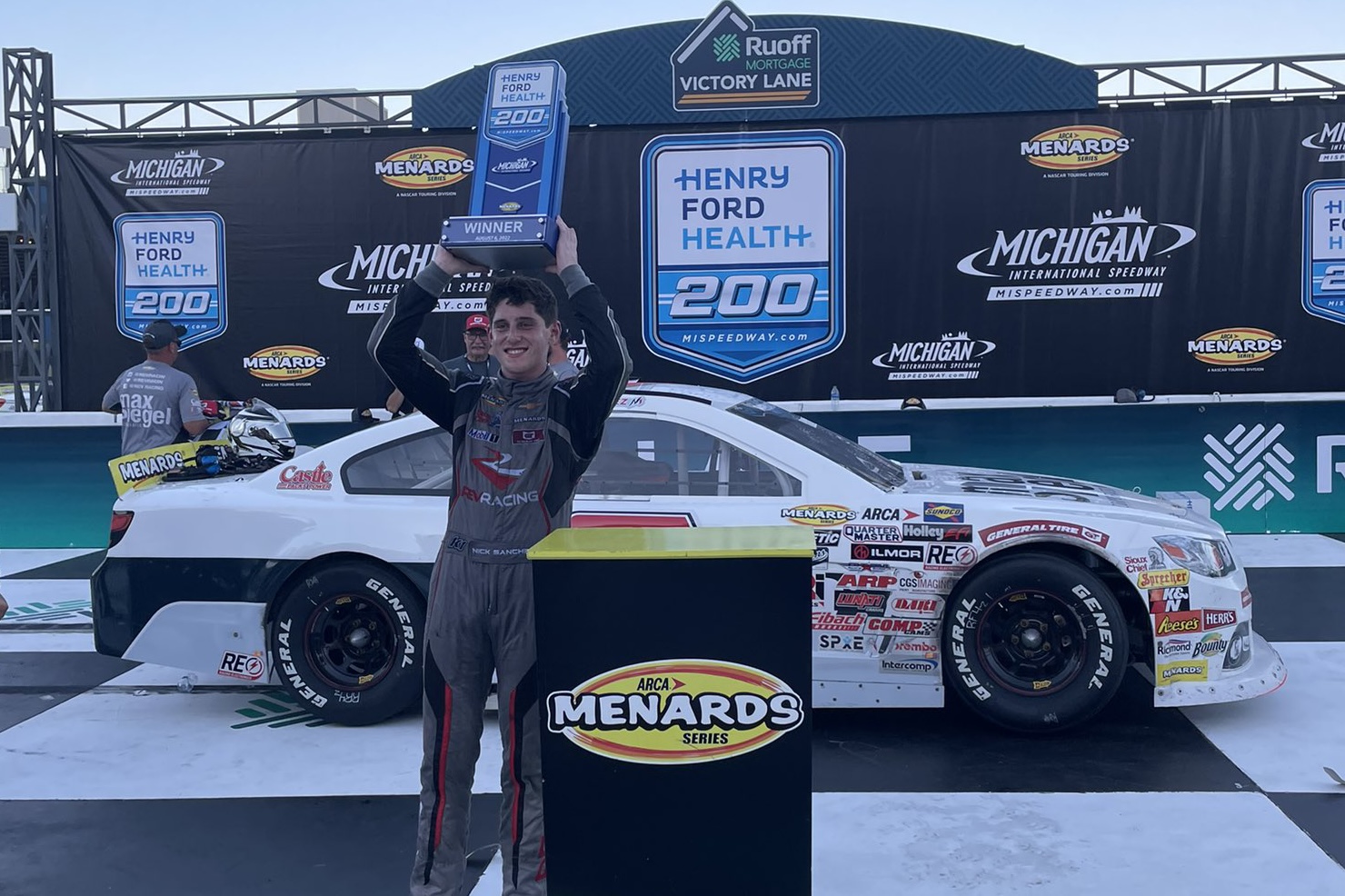 Nick Sanchez holds the Henry Ford Health 200 trophy above his head in victory lane at the Michigan International Speedway.